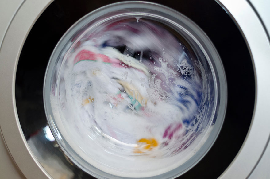 Wasmachine met witte zeep en gekleurde kleding draait achter een glazen deur met een zwarte en zilveren rand.