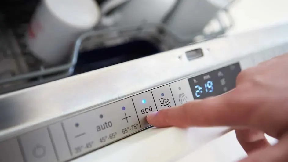A finger presses the eco button on a dishwasher. The display shows 2:19 and cups are in the top rack. No brand is visible.