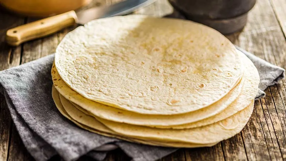 Une pile de tortillas sur un linge gris, avec un couteau et un mortier en arrière-plan sur une table en bois.