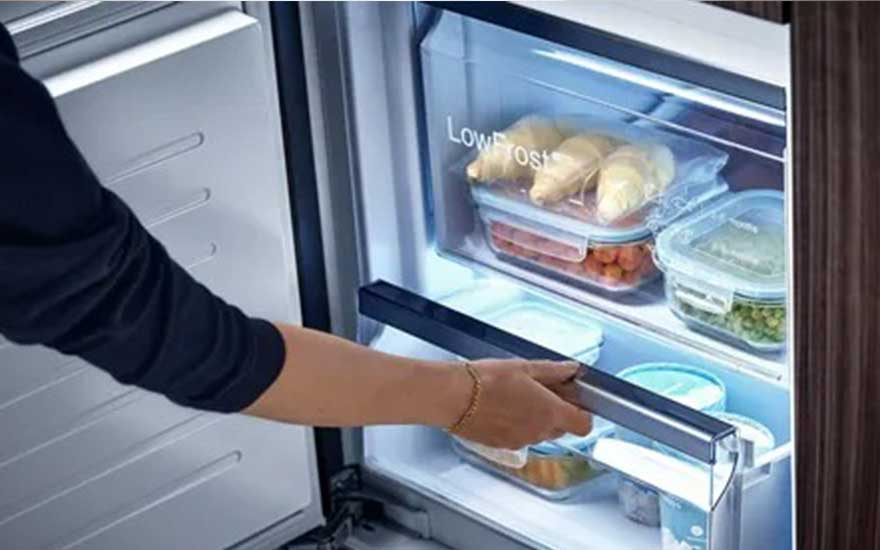 Une personne ouvre un réfrigérateur rempli de récipients transparents contenant divers aliments. L'étiquette "Low Frost" est visible.