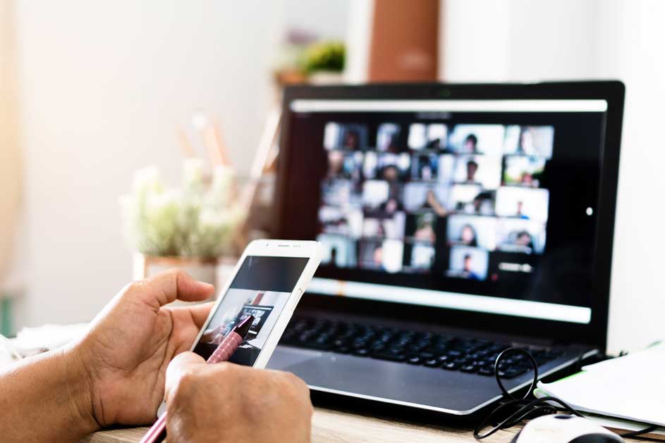 Een laptop en smartphone tonen een video call. Een hand houdt een stylus bij de smartphone. Op de achtergrond staat een plant.