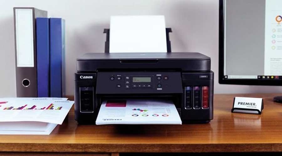 Canon inkttankprinter op een bureau, print kleurrijke grafieken; ordners links en monitor rechts.