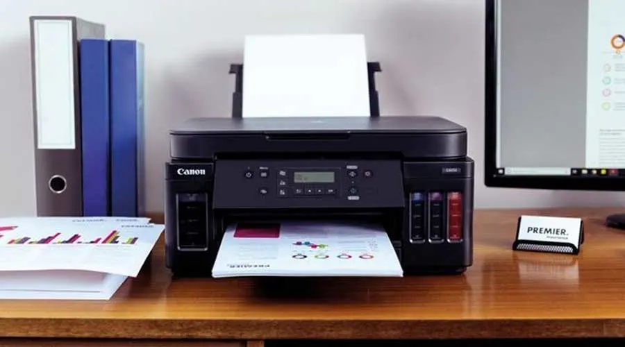 Canon inkttankprinter op een bureau, print kleurrijke grafieken; ordners links en monitor rechts.