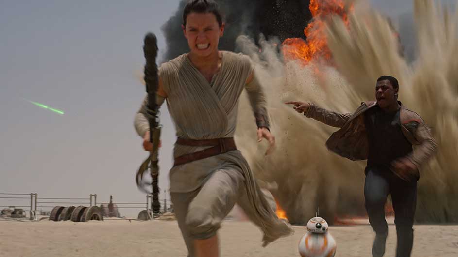 Rey court dans le désert avec un bâton. BB-8 et un autre coureur sont derrière, une explosion en arrière-plan.