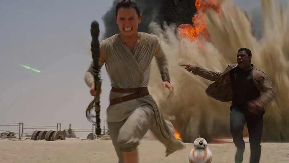 Rey court dans le désert avec un bâton. BB-8 et un autre coureur sont derrière, une explosion en arrière-plan.
