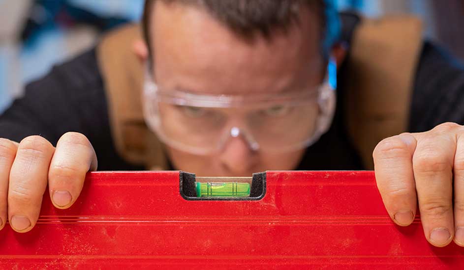 Un niveau rouge avec une bulle verte est tenu par des mains. Des lunettes de protection sont portées en arrière-plan.