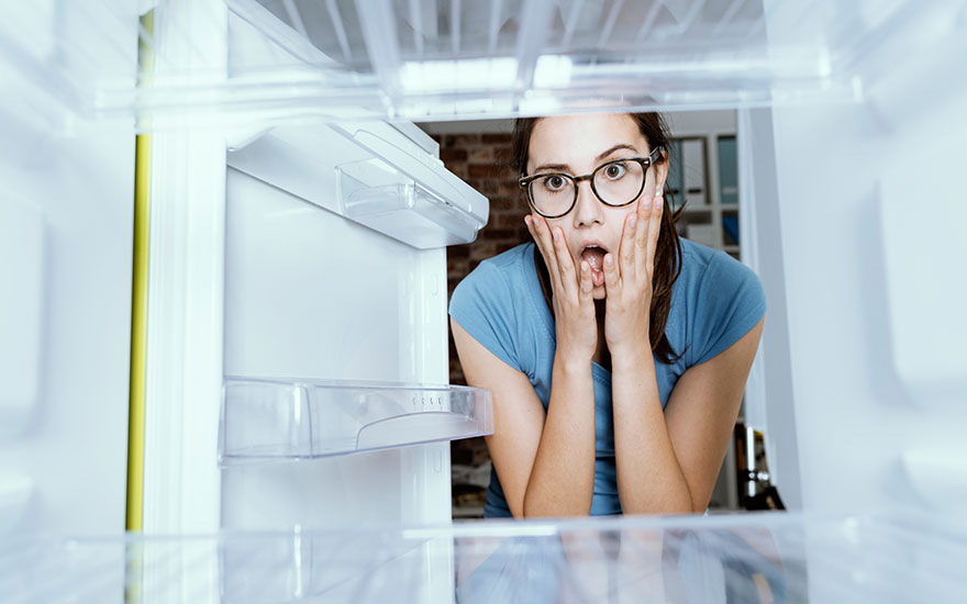 A nearly empty white refrigerator with clear shelves is in the foreground. A brick wall is visible in the background.