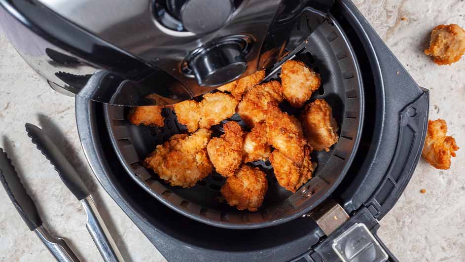 Een zwarte airfryer met goudbruine stukken gebakken kip. Een zilveren tang ligt ernaast op een gemarmerde ondergrond.