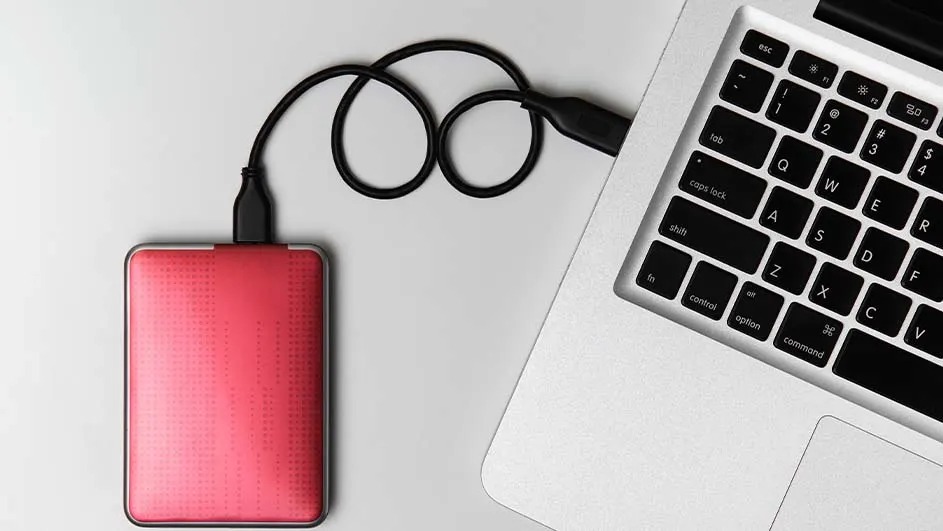 Een rode externe harde schijf is met een zwarte USB-kabel aangesloten op een zilveren laptop, geplaatst op een lichtgrijze ondergrond.