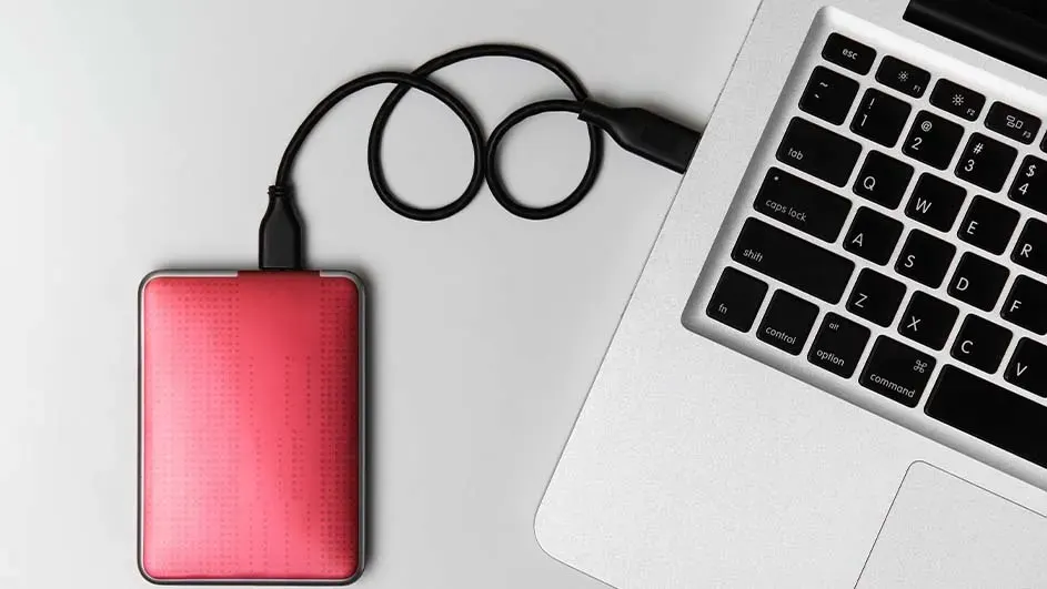 Een rode externe harde schijf is met een zwarte USB-kabel aangesloten op een zilveren laptop, geplaatst op een lichtgrijze ondergrond.
