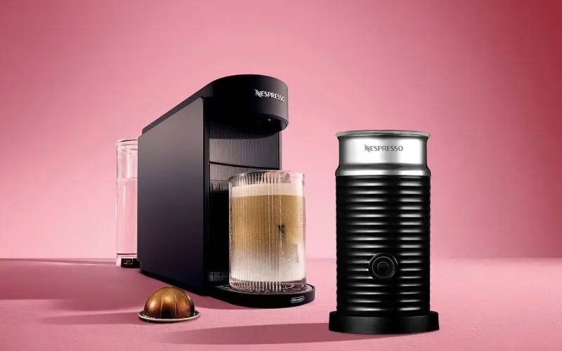 Een zwarte Nespresso-koffiemachine met een romige koffiedrank in een glazen beker, naast een zwarte Aeroccino-melkopschuimer en een goudkleurige capsule op een roze achtergrond.