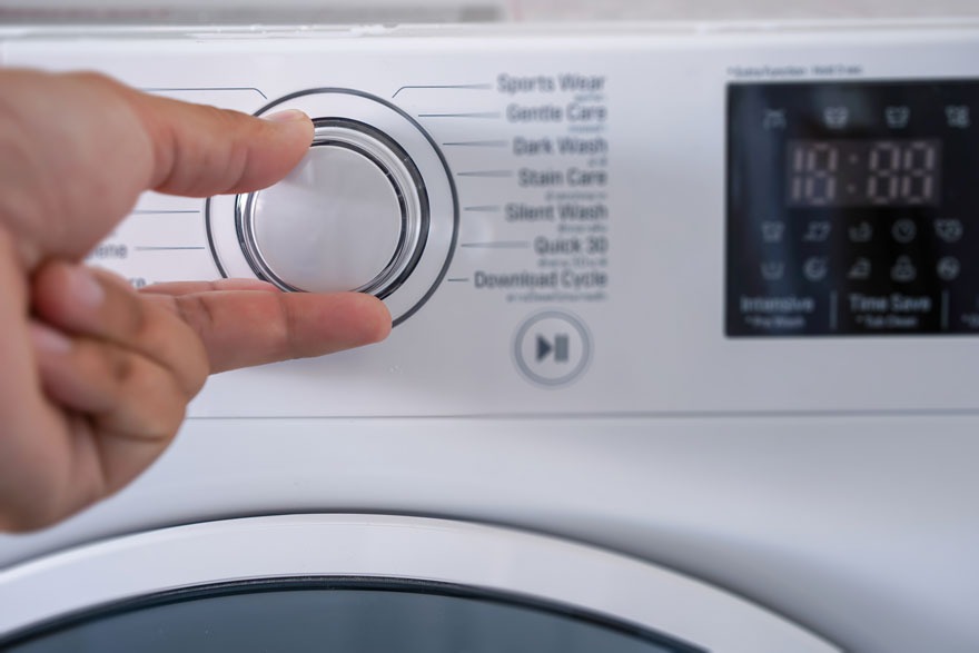 Une main ajuste le sélecteur de programme sur un lave-linge blanc avec écran numérique affichant l'heure.