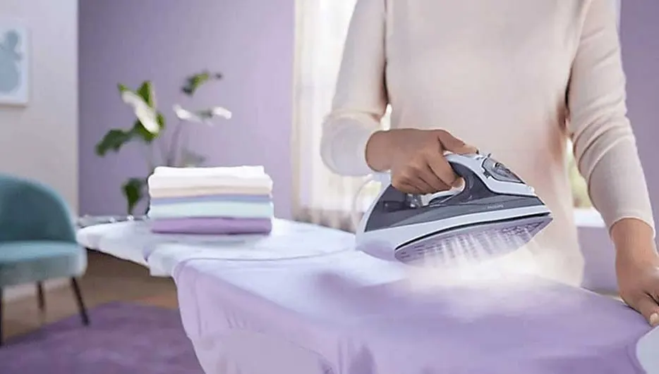 Une personne repasse un tissu violet sur une table à repasser. Un fer gris et blanc est utilisé. Des piles de linge sont visibles.