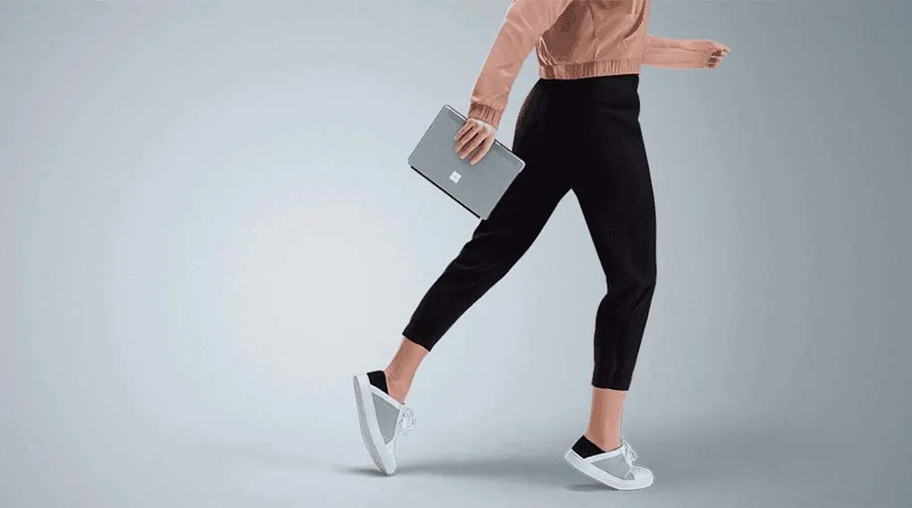 Een persoon in beweging houdt een grijze laptop vast, met zwarte broeken en witte schoenen tegen een grijze achtergrond.