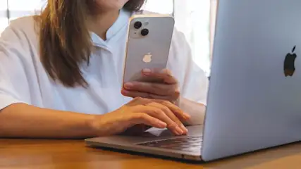Een laptop en een zilverkleurige telefoon staan op een houten tafel. De laptop is open en de telefoon wordt vastgehouden.