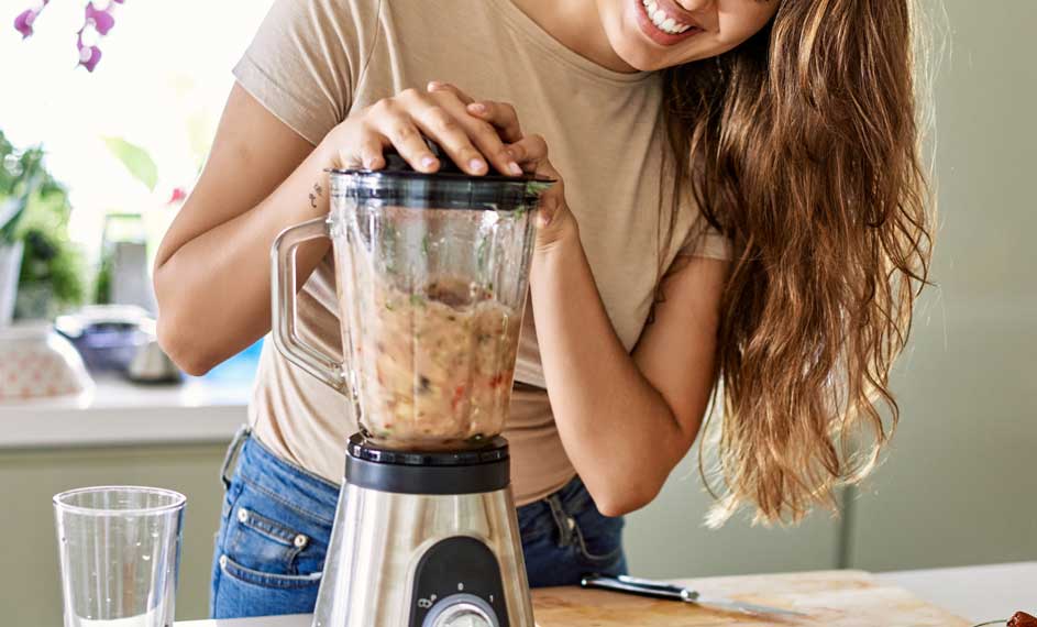 Een blender met een doorzichtige kan vol gemixt voedsel staat op een aanrecht. Een glas staat ernaast, klaar om gevuld te worden.