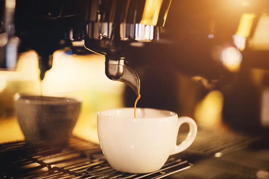 Een close-up van een koffiemachine die koffie in een witte mok tapt. Een andere mok staat op de achtergrond.