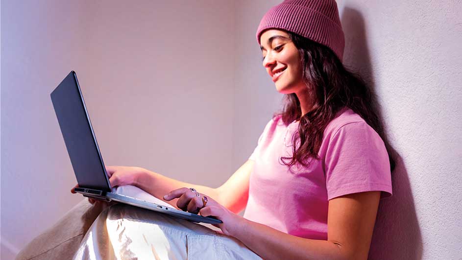 Glimlachende vrouw met roze muts gebruikt een laptop terwijl ze tegen een muur zit.