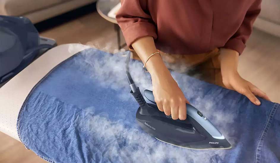 Une chemise en jean bleu est repassée sur une planche à repasser blanche avec un fer bleu et noir. De la vapeur s'échappe.