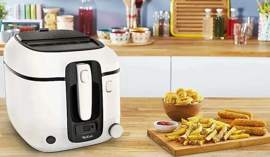 Een witte Tefal friteuse staat op het aanrecht naast een plank met frietjes, uienringen en snacks met saus.