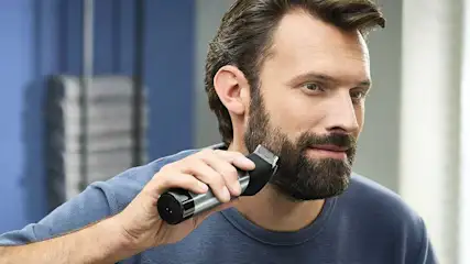 Een man met een blauw shirt trimt zijn baard met een elektrische trimmer in een moderne, lichtblauwe badkamer.