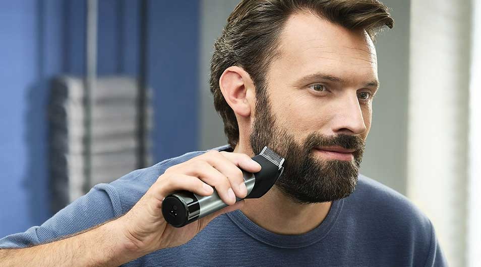 Un homme en t-shirt bleu taille sa barbe avec une tondeuse électrique dans une salle de bain moderne bleue.