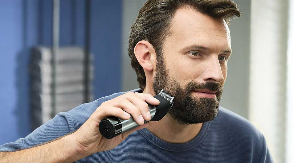 A man is using a black and silver electric razor on his beard. Background shows blue and white vertical lines.
