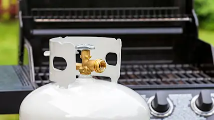 Bouteille de propane blanche avec raccord en laiton, devant un barbecue noir. L'arrière-plan est vert.