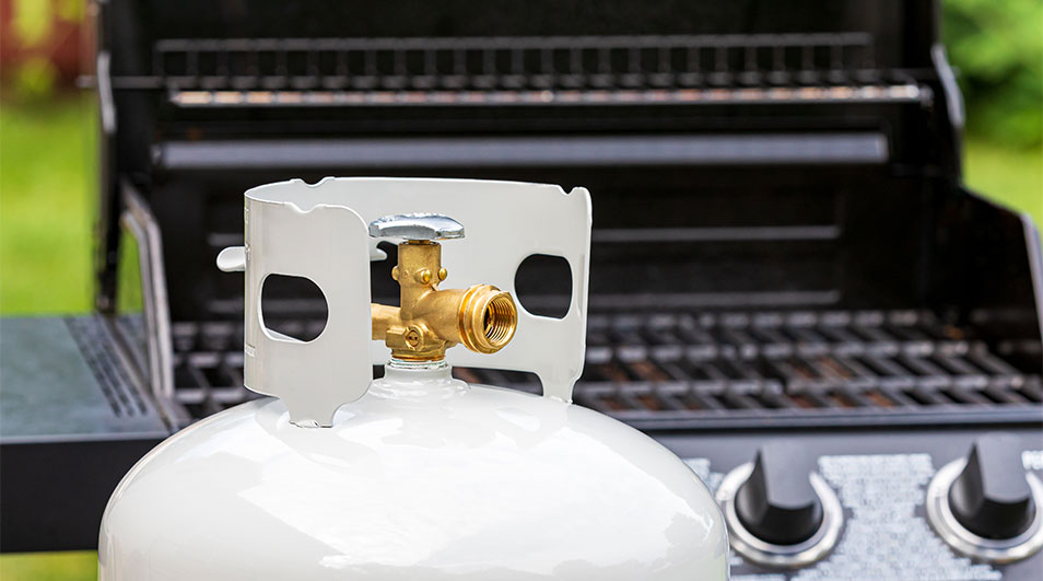 Bouteille de propane blanche avec raccord en laiton, devant un barbecue noir. L'arrière-plan est vert.