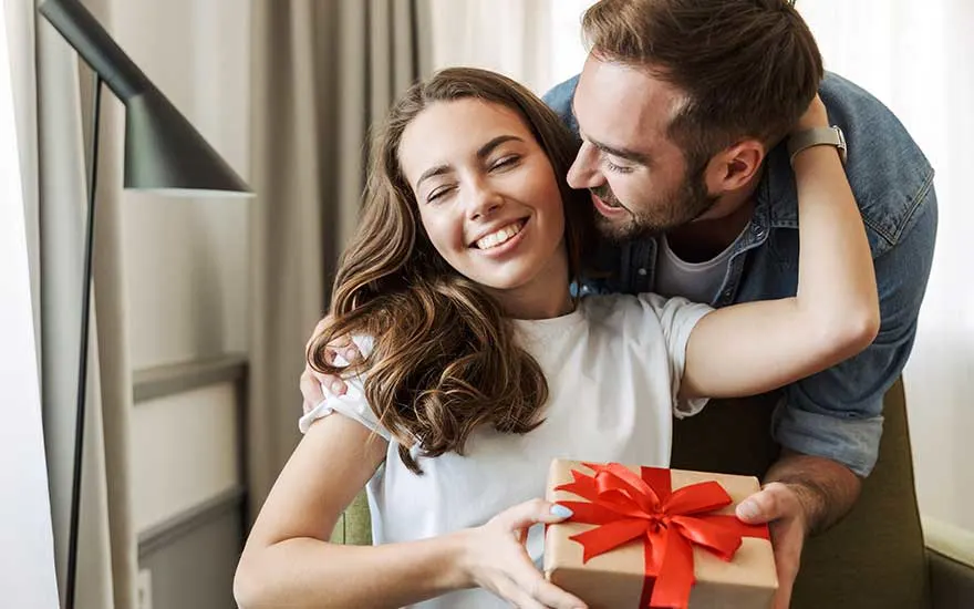 Une jeune femme sourit les yeux fermés en tenant un cadeau au ruban rouge, tandis que son partenaire la serre tendrement contre lui.