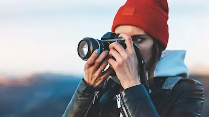 Une personne prend une photo avec un appareil photo noir devant un paysage flou. Elle porte un bonnet rouge et une veste en cuir noire.