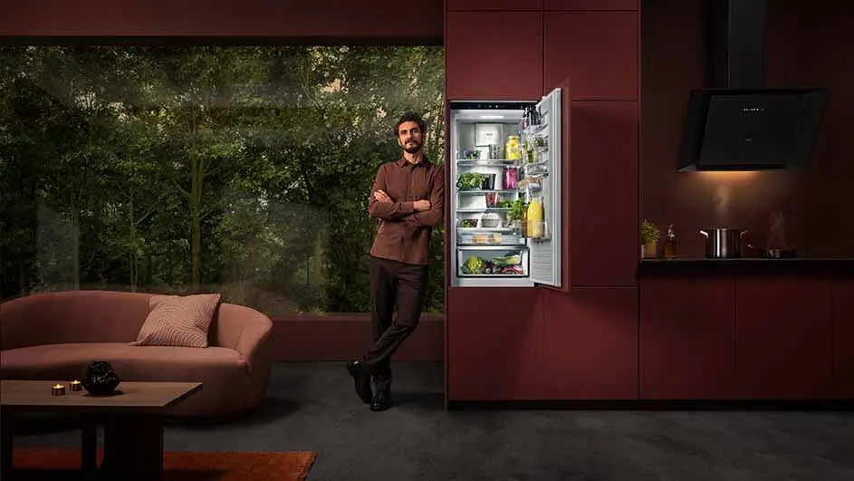 Woonkamer met een open koelkast, rode kasten, een grijze vloer en een raam met uitzicht op bomen.