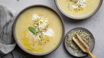 Deux bols de soupe jaune garnis de crème, graines et une feuille. Une assiette de graines avec une cuillère en bois.