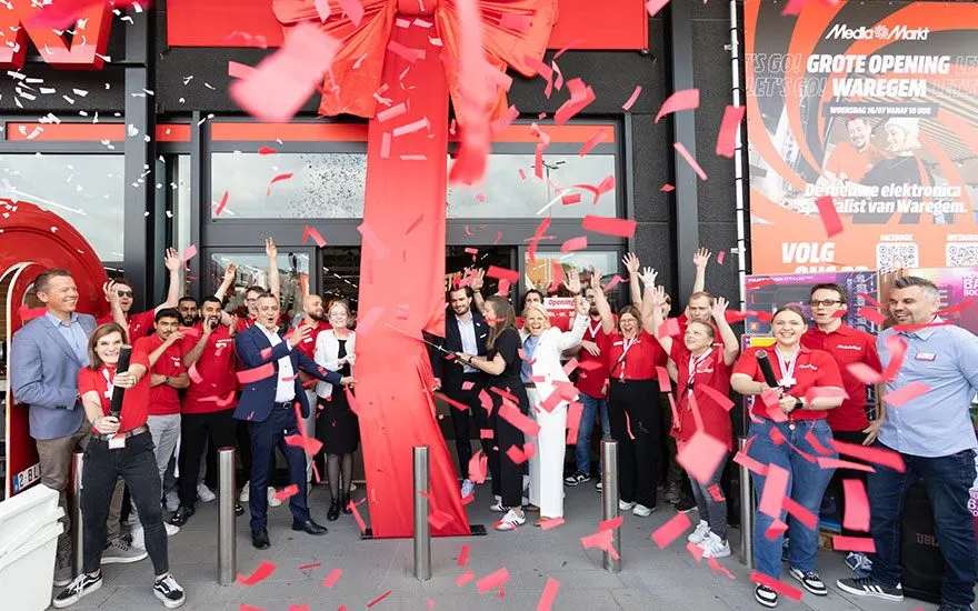 Ouverture de magasin MediaMarkt avec une équipe enthousiaste jetant des confettis devant l'entrée. Rubans rouges tombent.