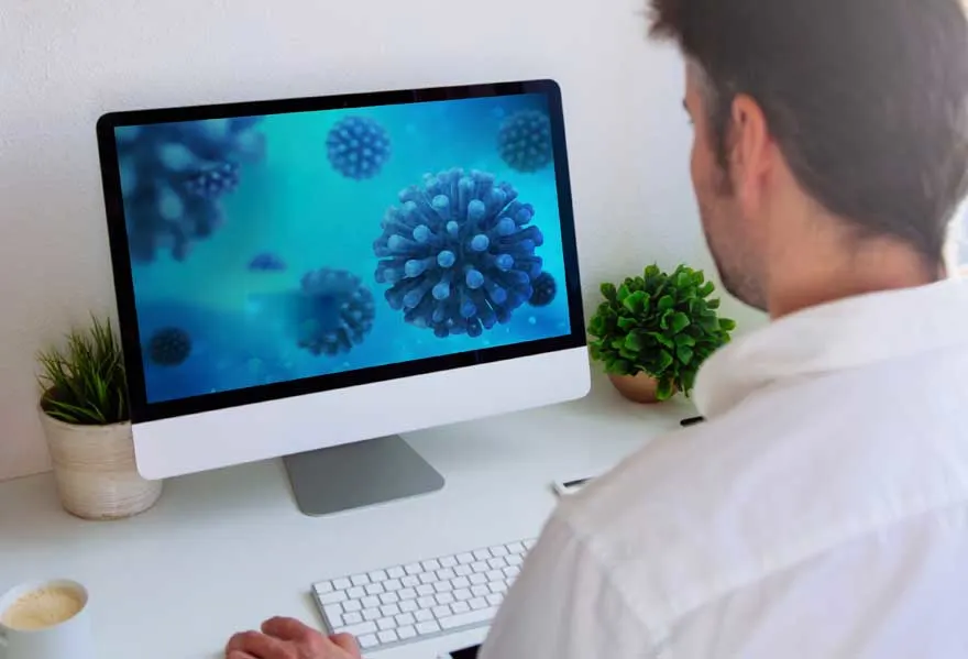 Bureau blanc avec un ordinateur affichant des images bleues de virus, un clavier, des plantes et une tasse.