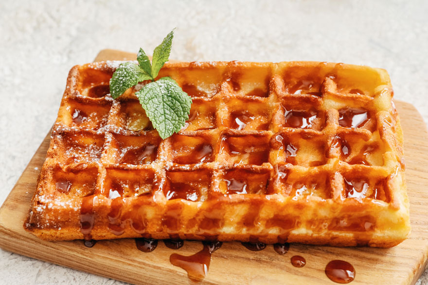 Gaufre dorée nappée de sirop sur une planche en bois. Quelques feuilles de menthe, et du sucre glace sur le côté.