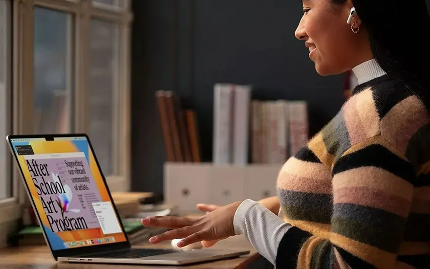 Personne en pull rayé utilise un MacBook Apple avec un programme créatif, livres en arrière-plan ; logo Apple bien visible.