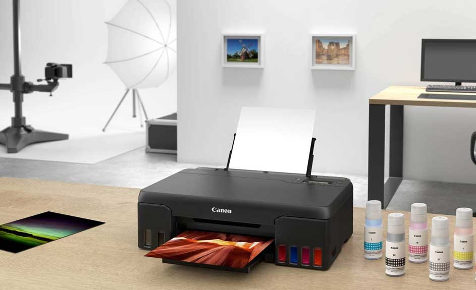 Een zwarte Canon printer print een foto op een beige tafel. Inktflessen en foto's liggen ernaast. Op de achtergrond staat een bureau en fotoapparatuur.
