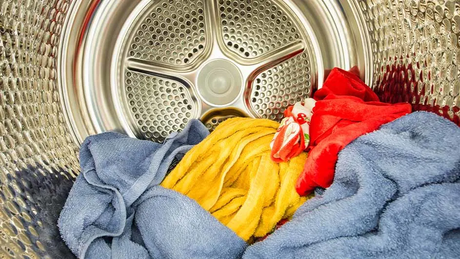 Inside a shiny metal dryer drum lie crumpled towels in blue, yellow and red, casually piled together and ready for a drying cycle.