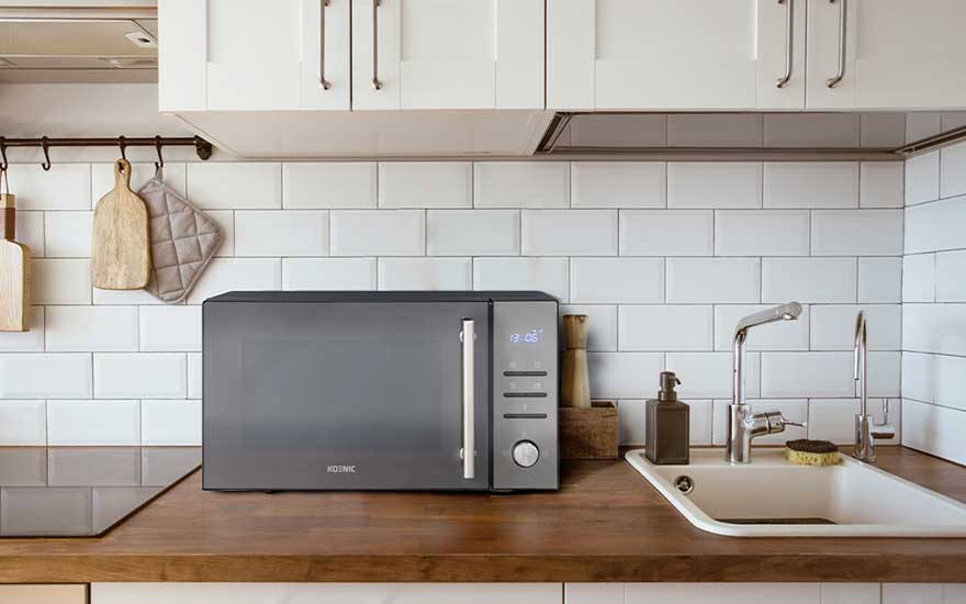 Modern kitchen featuring grey microwave on wooden worktop. White tiles, white cupboards and kitchen accessories in neutral tones.