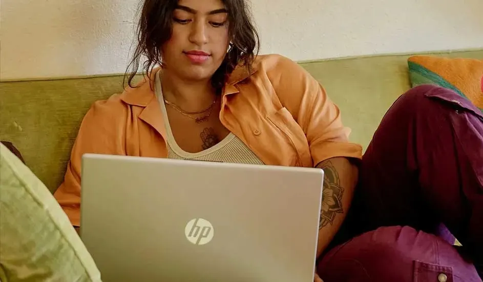 Person relaxing on green couch with open HP laptop on lap, wearing orange shirt and purple pants, calmly working from home.
