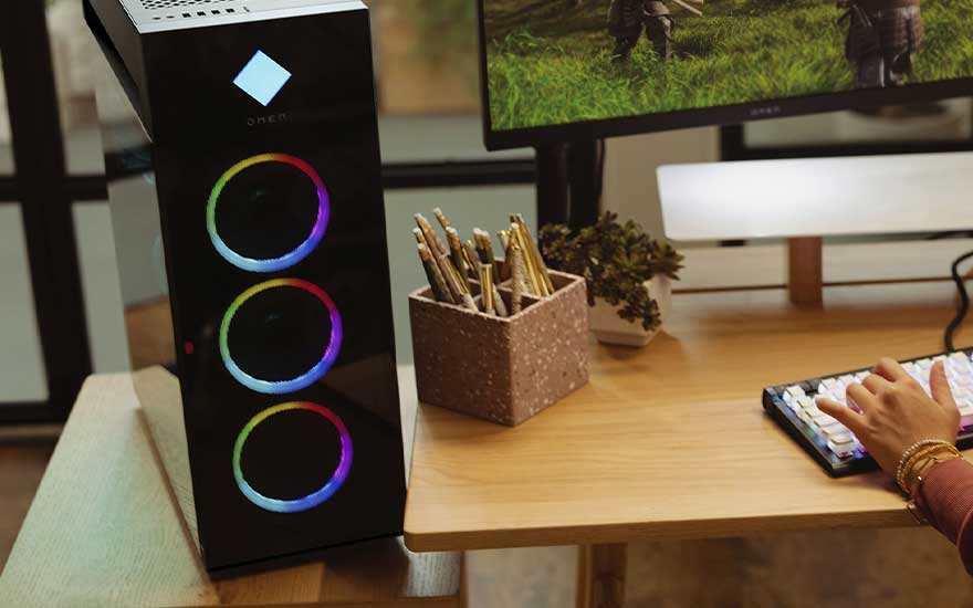 A desk with two monitors displaying a gradient of orange, pink, and blue. A keyboard, mouse, PC tower, and accessories are also visible.