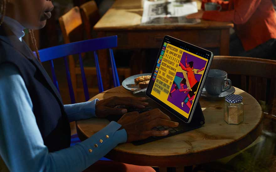 Sur une table en bois, un iPad affiche un texte coloré, à côté d'une tasse et d'un bocal. Une personne tape sur le clavier.