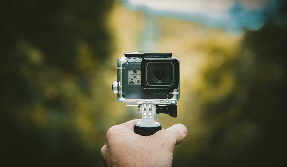 Een hand houdt een zwarte GoPro-camera in een doorzichtige behuizing tegen een vage achtergrond van groen en geel.