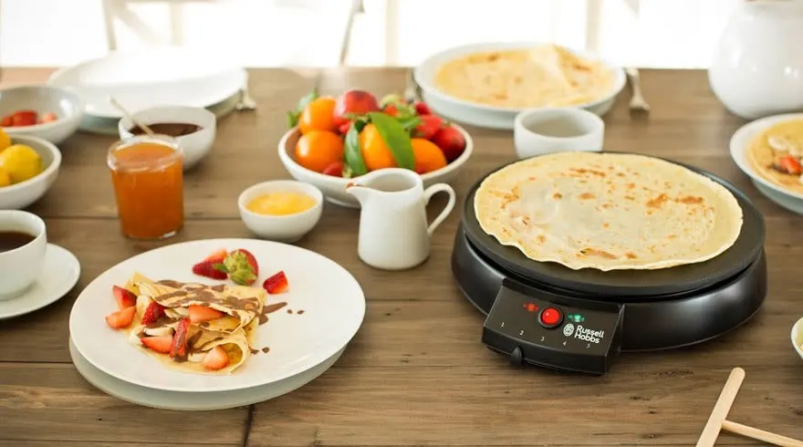 Table en bois avec crêpière, crêpes, fruits, café, jus et garnitures.