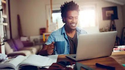 Un homme souriant travaille chez lui sur un ordinateur portable, une tasse bleue à la main, avec papiers et livres sur la table.