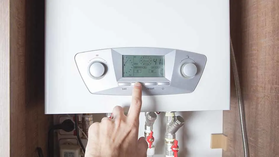 Un doigt appuie sur un bouton d'une chaudière blanche avec un écran affichant des chiffres, au-dessus de tuyaux métalliques.
