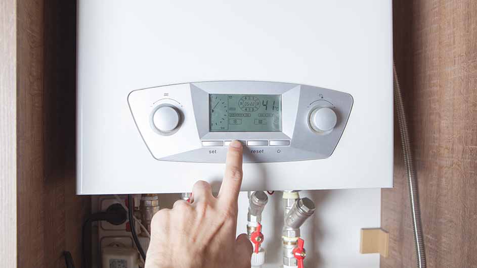 Een witte verwarmingsketel aan de muur met een digitaal display en rode kraantjes. Een vinger drukt op de resetknop.