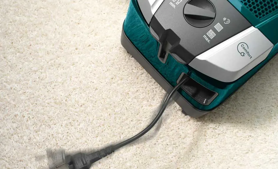Aspirateur turquoise et gris foncé sur un tapis texturé beige avec un câble noir branché.