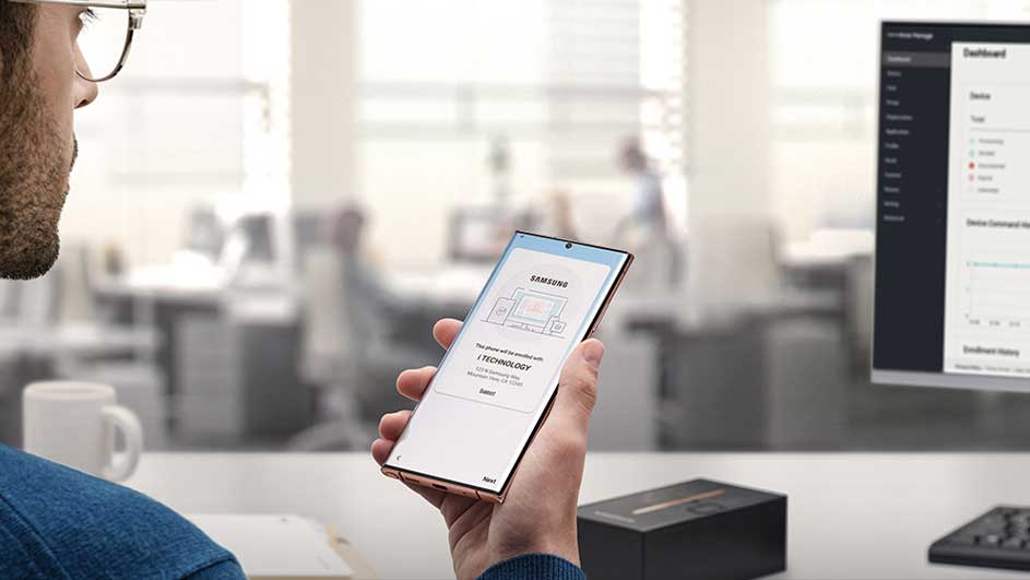Un téléphone Samsung est tenu devant un écran d'ordinateur dans un bureau moderne et lumineux.